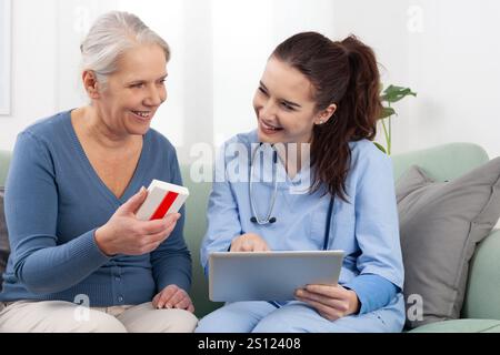 Eine junge freundliche Krankenschwester oder Ärztin lächelt, während sie einer älteren Patientin die Medikamente auf einem digitalen Tablet erklärt und zeigt. Zu Hause, in einem Arzt Stockfoto