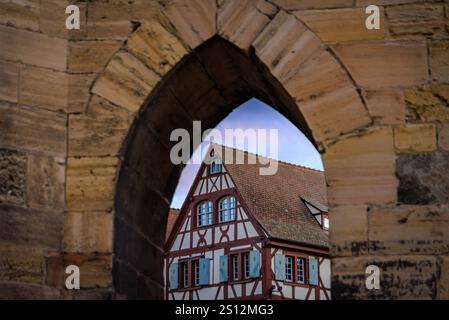 Traditionelles altes, halbes Fachwerkhaus mit roten Balken an der weißen Fassade, blaue Fensterläden, die bei Sonnenuntergang durch einen Steinbogen in Colmar, Frankreich, gesehen werden Stockfoto