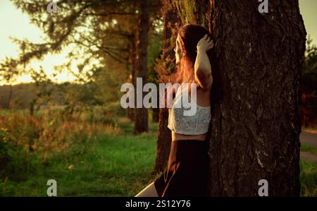 Schöne Frau, die den Rücken zur Eiche steht und auf den erstaunlichen Sommer hellen Sonnenuntergang in den orange Sonnenstrahlen auf dem Feld mit Bäumen und Grün blickt Stockfoto
