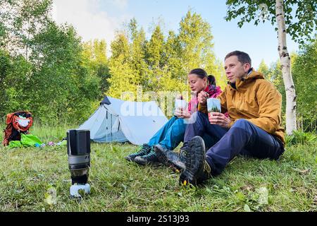 Ein Paar sitzt auf Gras neben dem Zelt im üppigen Wald und genießt Mahlzeiten aus verpackten Beuteln. In Outdoor-Ausrüstung gekleidet, mit tragbarem Campingofen und Rucksack in der Nähe. Grüne Bäume und Laub auf dem Hintergrund. Stockfoto