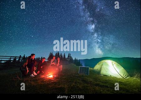 Eine Gruppe von Wanderfreunden ruht am Lagerfeuer neben einem beleuchteten Zelt und einem Solarpanel unter sternenüberzogenem Himmel. Die Milchstraße leuchtet hell über den Bergen und schafft eine magische Atmosphäre in der Wildnis der Berge. Stockfoto