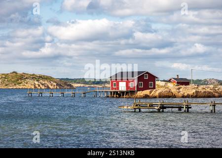 Meereslandschaft in der Provinz Bohuslän, Schweden, mit einem rot bemalten Bootshaus an der felsigen Küste mit einem hölzernen Pier unter stürmischem Himmel an einem sonnigen Tag. Stockfoto