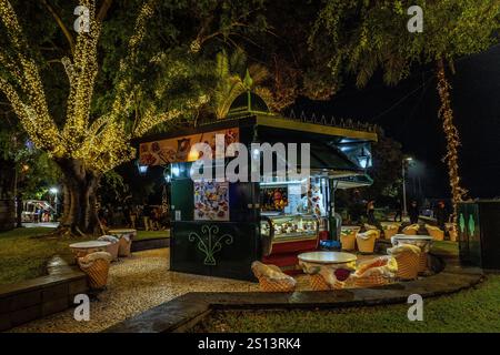 Ein Café-Kiosk im Freien, beleuchtet bei Nacht, in einem üppigen Park mit dekorierten Bäumen, schafft eine gemütliche und einladende Atmosphäre für Entspannung und Geselligkeit Stockfoto