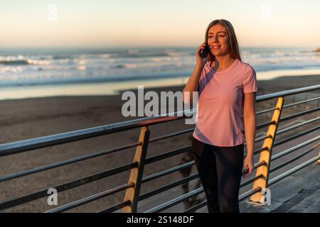 Die junge Sportlerin telefoniert, während sie den Sonnenuntergang am Strand genießt, Sportbekleidung trägt und sich nach dem Inline-Skating-Training entspannt fühlt Stockfoto