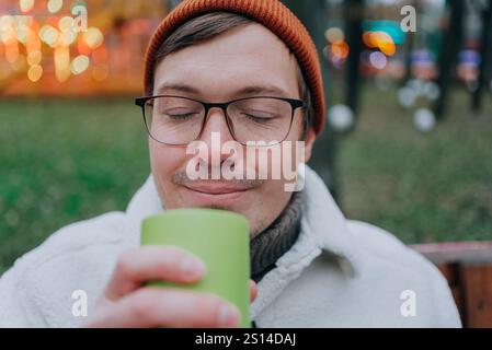 Ein junger Mann mit Brille und Wollmütze genießt auf einem weihnachtsmarkt im Freien ein warmes Getränk und genießt die festliche Atmosphäre und den Duft seines Getränks Stockfoto