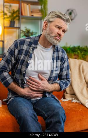 Ein älterer Mann sitzt auf der Couch und fühlt plötzlich starke Bauchschmerzen, Gastritis-Problem. Mann mittleren Alters mit Symptom Vergiftung Durchfall Verdauungsstörungen peptisches Ulkus Pankreatitis zu Hause. Vertikal Stockfoto