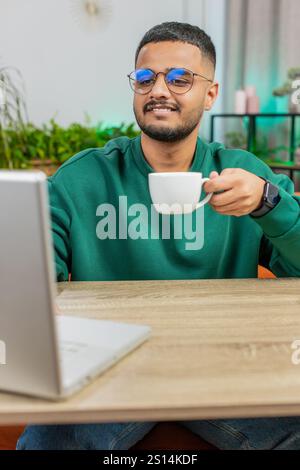 Attraktiver lächelnder indischer Mann, der am Laptop arbeitet und eine Tasse warmen heißen Kaffees trinkt, sitzt morgens am Tisch, am Arbeitsplatz, im Home Office. Ein Araber genießt es, sich zu entspannen und eine Pause zu machen. Vertikal Stockfoto