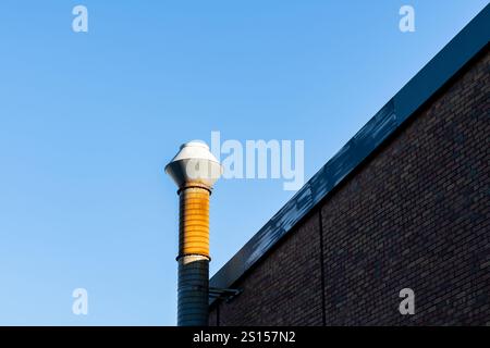 Ein zylindrischer Rauchstapel erhebt sich aus einem Ziegelgebäude und reicht nach oben in einen klaren blauen Himmel. Die Ziegelfassade des Gebäudes steht im Kontrast zu den hellen s Stockfoto