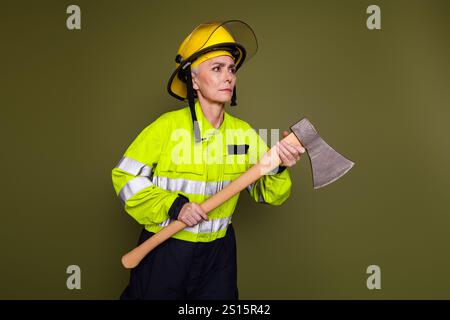 Reife weibliche Feuerwehrfrau in gut sichtbarer Schutzuniform, die eine Axt vor khakifarbenem Hintergrund hält Stockfoto