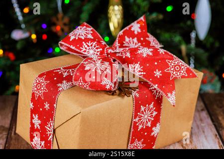 Wunderschön gewickeltes Geschenk mit rotem Schneeflockenband sitzt auf Holzoberfläche neben Weihnachtsgeschenken, frohe Weihnachten bis hell erleuchteter Weihnachtsbaum. Stockfoto