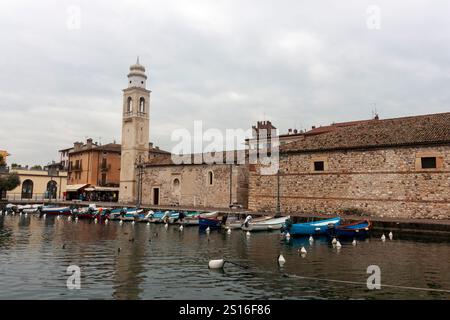 LAZISE, ITALIEN - 25. OKTOBER 2024: Ein malerischer Blick auf den alten Hafen von Lazise, mit farbenfrohen Booten, die entlang der Uferpromenade und dem Kirchturm i vertäut sind Stockfoto