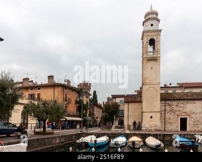 LAZISE, ITALIEN - 25. OKTOBER 2024: Ein malerischer Blick auf den alten Hafen von Lazise, mit farbenfrohen Booten, die entlang der Uferpromenade und dem Kirchturm i vertäut sind Stockfoto