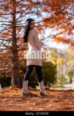 Junge Frau in beigem Kleid und Cowboystiefeln, die im Herbst in einem Park posiert Stockfoto