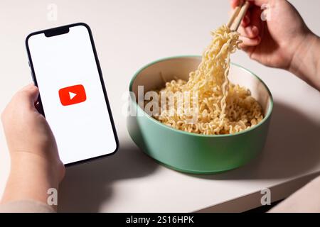 Frau, die die Youtube-App auf dem Smartphone nutzt, während sie zu Hause Nudeln isst. YouTube-Logo auf dem Bildschirm. Essen mit Social-Media-Konzept Stockfoto