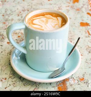 Ein flacher weißer Kaffee in einer hellblauen Tasse und Untertasse mit Teelöffel auf einer abgenutzten, rustikalen Tischplatte, die in hellen Blau- und Orangetönen harmoniert. Stockfoto