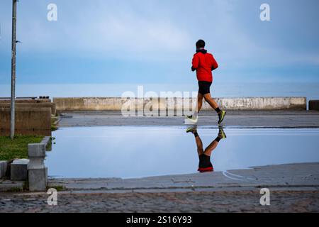 Ein Mann in einer roten Jacke läuft auf einem nassen Bürgersteig. Das Wasser reflektiert den Himmel und das Spiegelbild des Mannes. Die Szene ist friedlich und ruhig, mit t Stockfoto