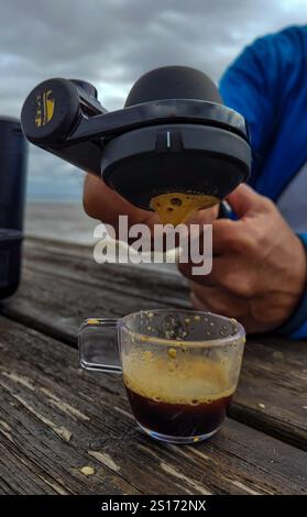 Die Hände der Herren halten die Reisekaffeemaschine Handpresso, aus der ein duftender frischer, kräftiger Espresso mit Schaum in eine Tasse auf einem Holztisch fließt. Stockfoto