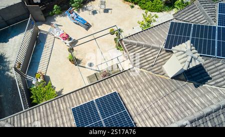 Eine Luftaufnahme zeigt zwei Frauen, die sich in einem sonnigen Hinterhof sonnen, während auf dem Dach Solarpaneele installiert sind. Die Szene zeigt die umweltfreundlichen und Stockfoto