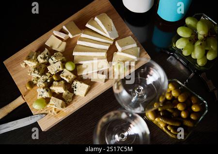 Ein Holzbrett mit verschiedenen Käsesorten, frischen grünen Trauben und Oliven steht auf einem Tisch. In der Nähe gibt es zwei leere Weingläser, eine Flasche und Platz Stockfoto