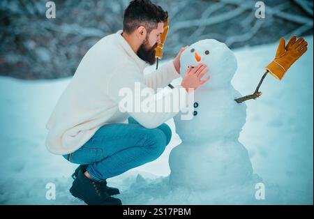 Schneemann und lustiger bärtiger Mann im Schnee. Mann, der mit Snowman spielt - isoliert auf Schnee Hintergrund. Fröhlich hübscher junger Hipster, der Spaß mit Schnee hat Stockfoto