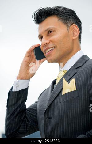 Lächelnder, elegant gekleideter Geschäftsmann aus Asien/dem Nahen Osten, der ein Mobiltelefon am Ohr hält, in einer Stadt im Freien. Stockfoto