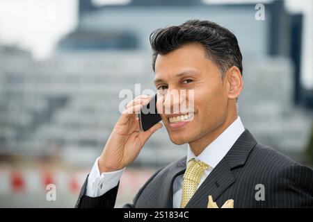 Lächelnder, elegant gekleideter Geschäftsmann aus Asien/dem Nahen Osten, der ein Mobiltelefon am Ohr hält, in einer Stadt im Freien. Stockfoto