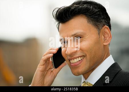 Lächelnder, elegant gekleideter Geschäftsmann aus Asien/dem Nahen Osten, der ein Mobiltelefon am Ohr hält, in einer Stadt im Freien. Stockfoto