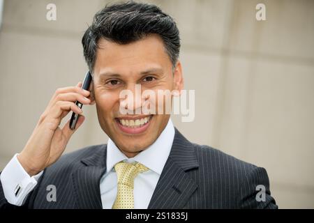 Lächelnder, elegant gekleideter Geschäftsmann aus Asien/dem Nahen Osten, der ein Mobiltelefon am Ohr hält, in einer Stadt im Freien. Stockfoto