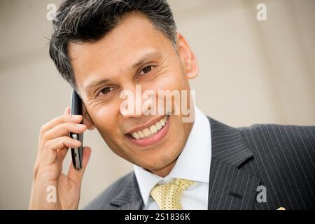 Lächelnder, elegant gekleideter Geschäftsmann aus Asien/dem Nahen Osten, der ein Mobiltelefon am Ohr hält, in einer Stadt im Freien. Stockfoto