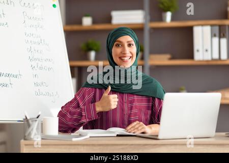 Islamischer Lehrer im Kopftuch, der am Schreibtisch sitzt und den Daumen nach oben zeigt Stockfoto