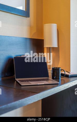 Ein Laptop auf dem Schreibtisch des Hotels, neben einem schwarzen Becher, einem Notizblock und einer kleinen Tischlampe. Stockfoto