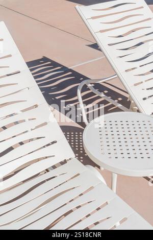 Weiße Liegestühle mit Tisch auf der Poolterrasse mit Schatten und Wellenmuster an sonnigen Tagen. Die Stühle werfen gemusterte Schatten. Stockfoto