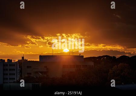 Die untergehende Sonne strahlt sanfte Strahlen von leuchtenden orangefarbenen Strahlen aus, die sich am Abendhimmel ablichten, während sie hinter einer Wolkenlinie absinkt, die den Horizont bedeckt. Stockfoto