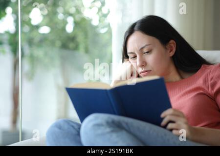 Gelangweilte asiatische Frau, die Papierbuch liest und zu Hause auf einer Couch sitzt Stockfoto