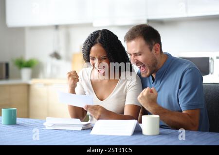 Aufregendes interrassisches Paar, das Rechnungen prüft und Erfolg in der Küche zu Hause feiert Stockfoto