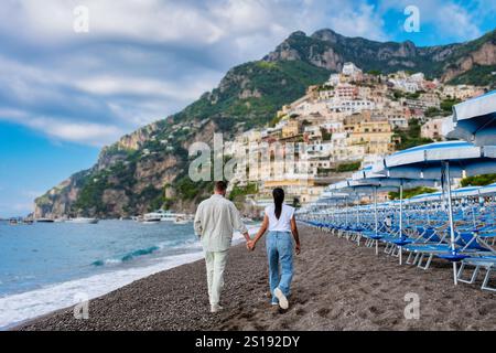 Ein paar spazieren entlang eines Sandstrandes, Hand in Hand, mit bunten Häusern, die sich an den Amalfi-Klippen über ihnen Klammern. Die ruhigen Wellen schlängeln sanft am Ufer unter einem dramatischen Himmel. Positano Italien Stockfoto