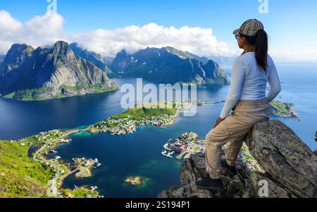 Asiatische Frauen mit einer atemberaubenden aussicht von einem felsigen Gipfel aus aufgenommen, die die atemberaubenden Lofoten-Inseln umgeben von glitzerndem Wasser unter einem klaren blauen Himmel zeigt. Reinebringen Lofoten Norwegen Stockfoto
