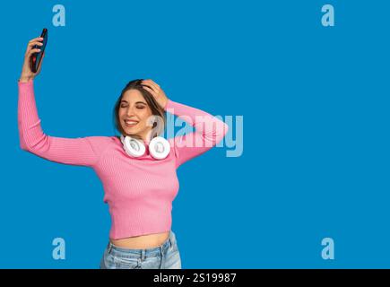 Glückliche junge Frau mit Kopfhörern um den Hals, das Smartphone hält und Musik hört, tanzt, isoliert auf blauem Studiohintergrund Stockfoto