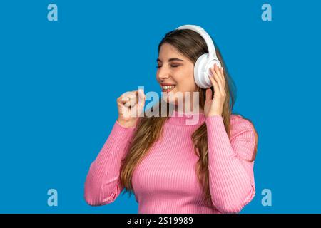 Glückliche junge Frau, die Musik mit kabellosen Kopfhörern hört, auf blauem Hintergrund tanzt und singt Stockfoto