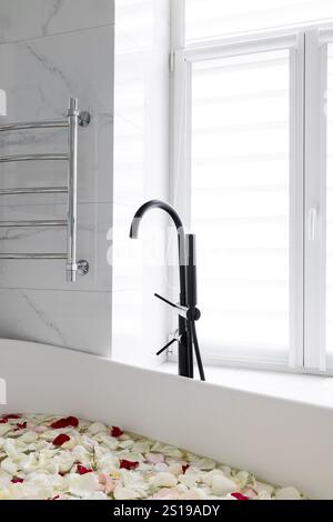 Moderne Badewanne mit elegantem schwarzen Wasserhahn, umgeben von weichen Rosenblättern Stockfoto