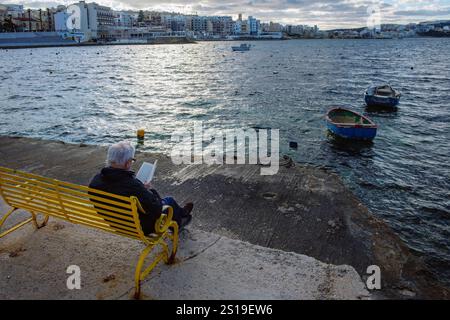 Ein Mann liest ein Buch an einem Winternachmittag am Hafen in St. Paul's Bay, Malta Stockfoto