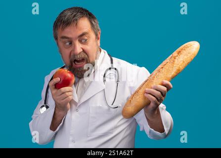 Ein Mann in einem weißen Labormantel und Stethoskop hält einen roten Apfel in der einen Hand, während er in der anderen aufgeregt auf ein langes Baguette blickt. Stockfoto