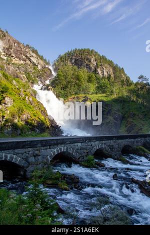 Latefossen, ein mächtiger Wasserfall, stürzt sich in felsige Berge, umgeben von üppigem Grün. Eine malerische Steinbrücke überspannt den Fluss. Stockfoto