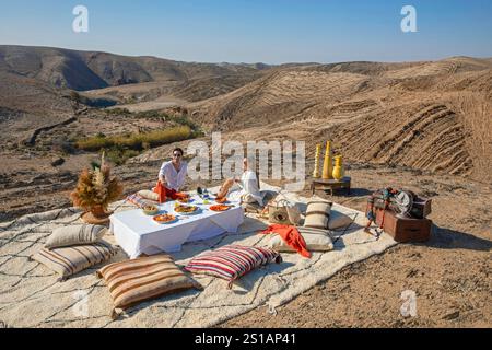 Marokko; Marrakeck, Agafay, Anara Camp, junge Paare, die auf Berberteppichen inmitten trockener Hügel picknicken Stockfoto