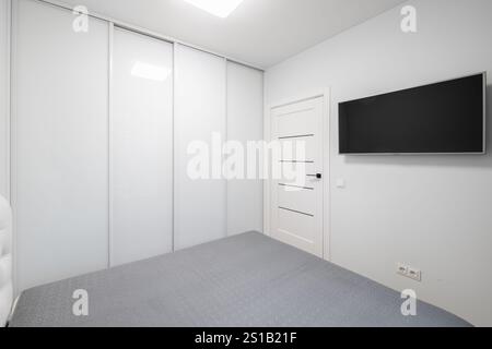 Minimalistischer Wohnzimmerinnenraum. Kingsize-Doppelbett, Kleiderschrank. Weiße Wände und Decken. Graue Decke auf dem Bett. LED-Leuchte. TV-Bildschirm Stockfoto
