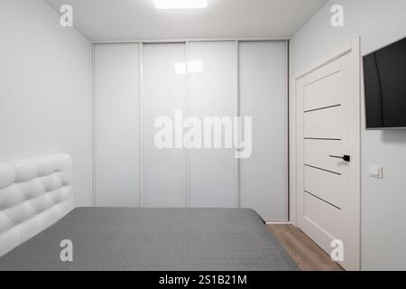 Minimalistischer Wohnzimmerinnenraum. Kingsize-Doppelbett, Kleiderschrank. Weiße Wände und Decken. Graue Decke auf dem Bett. LED-Leuchte. TV-Bildschirm Stockfoto