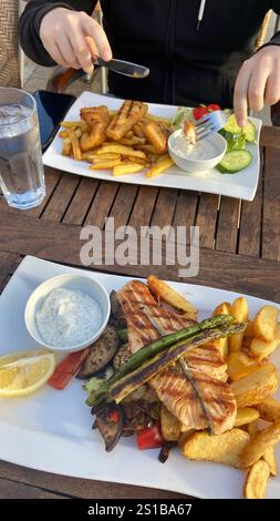 Speisen im Freien, wobei die Hände gegrillter Lachs, Gemüse und Kartoffelscheiben sowie gebratener Fisch und Chips zuschneiden Stockfoto