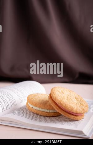 Fotos Sie köstliche Sandwichkekse mit Creme-Füllung auf offenem Buch vor luxuriösem braunem Stoffhintergrund für ein gemütliches Leseerlebnis und Stockfoto