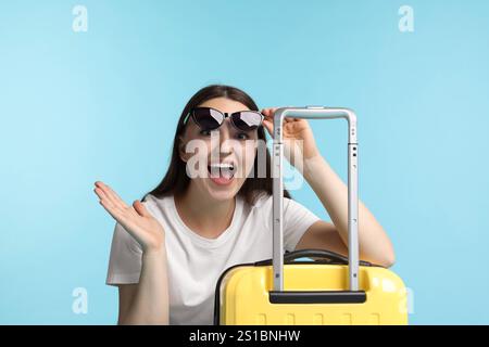 Frau mit Koffer, die Sonnenbrille auf hellblauem Hintergrund trägt Stockfoto