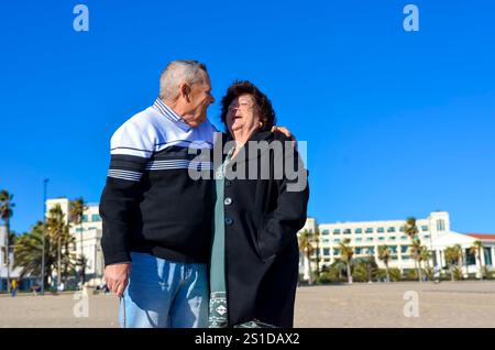 Seniorenpaar, das einen sonnigen Tag am Strand genießt, umgeht und lächelt Stockfoto